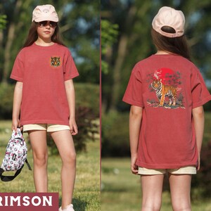 Op de afbeelding: Een rood T-shirt met een tijgerprint op de voorkant en een tijgerprint met een rode zon en bomen op de achterkant. Het shirt wordt gedragen door iemand met een roze baseballpet.