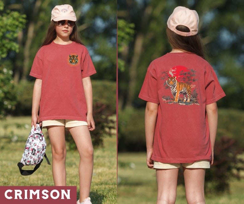 Op de afbeelding: Een rood T-shirt met een tijgerprint op de voorkant en een tijgerprint met een rode zon en bomen op de achterkant. Het shirt wordt gedragen door iemand met een roze baseballpet.