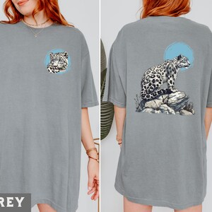 May include: A grey t-shirt featuring a graphic of a snow leopard sitting on a rock. The snow leopard is white with black spots and is looking to the right. The graphic is on the back of the shirt.