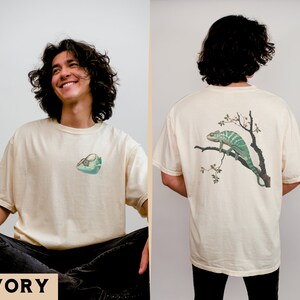 May include: A cream colored t-shirt with a green chameleon on a branch with white flowers on the back. The front of the shirt has a small graphic of a chameleon on a branch. The text "IVORY" is at the bottom of the image.