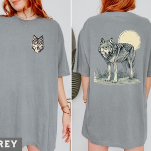 May include: Two grey t-shirts with a wolf graphic. One shirt features a wolf head graphic, while the other shirt features a wolf howling at the moon.