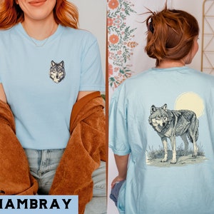 May include: Two light blue t-shirts with a wolf graphic. The shirt on the left has a wolf's head graphic, while the shirt on the right has a wolf howling at the moon graphic. The word "CHAMBRAY" is printed in black at the bottom of the image.
