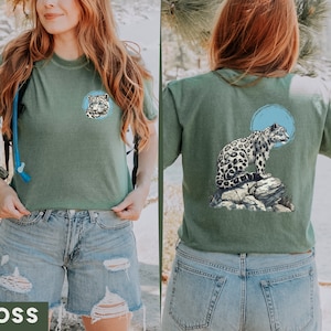 May include: A green t-shirt featuring a snow leopard graphic on the back and a snow leopard head on the front. The shirt is being modelled by a person wearing blue denim shorts.