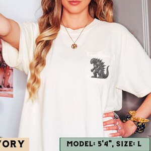 May include: Ivory-colored t-shirt with a pocket featuring a Godzilla graphic. The shirt is a size L. The model is 5'4". The shirt is a relaxed fit and has short sleeves.