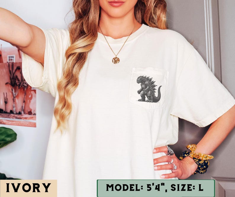 May include: Ivory-colored t-shirt with a pocket featuring a Godzilla graphic. The shirt is a size L. The model is 5'4". The shirt is a relaxed fit and has short sleeves.