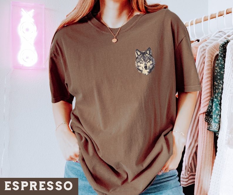 May include: A brown t-shirt with a wolf graphic on the left chest. The wolf is embroidered in brown, black, and white.