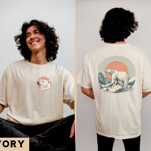 May include: A cream colored t-shirt with a polar bear graphic on the front and a larger polar bear graphic on the back. The back graphic features a polar bear walking on ice with mountains and a sunset in the background. The text "IVORY" is printed in black at the bottom of the image.