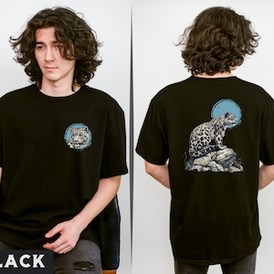 May include: Black t-shirt featuring a graphic of a snow leopard on a rock with a blue circle behind it. The graphic is on the back of the shirt. The shirt is being modelled by a person.