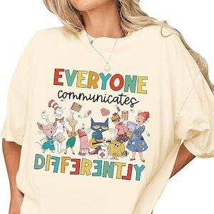 Puede incluir: Camiseta color crema con el texto "EVERYONE communicates DIFFERENTLY" en letras de bloque coloridas. La camiseta presenta una ilustración caprichosa de varios personajes de dibujos animados, incluidos animales y personas.