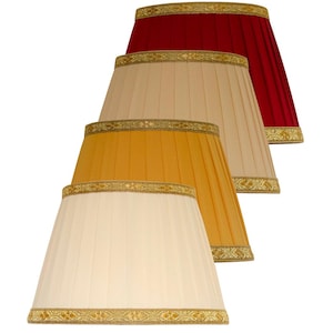 May include: A set of four pleated lamp shades in cream, gold, beige, and deep red. Each shade features a tapered bell shape with a decorative gold-toned woven trim along the top and bottom edges, providing a consistent design across all colors.
