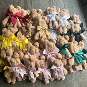 May include: A collection of tan towel bears, each shaped from a folded washcloth and adorned with a satin ribbon bow in various colors including burgundy, white, gold, black, yellow, pink, and dark green, arranged in a group on a flat surface.