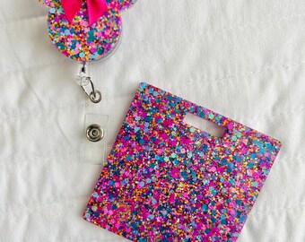 May include: A colorful glitter badge holder with a pink bow and a matching glitter badge insert. The badge holder is shaped like Mickey Mouse ears.