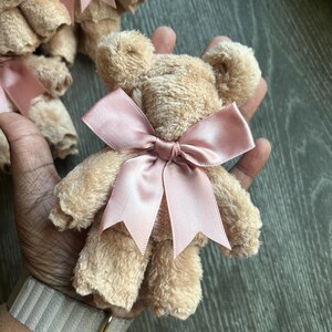May include: A small teddy bear crafted from beige, textured fabric, featuring a smooth, dusty pink satin ribbon tied into a bow around its neck. The bear has rounded ears and a soft, plush appearance, with additional similar bears visible in the background.