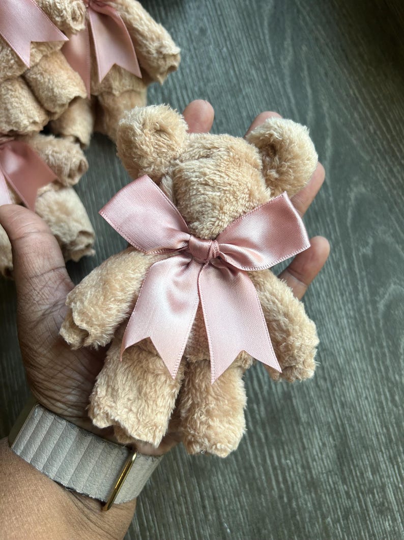 May include: A small teddy bear crafted from beige, textured fabric, featuring a smooth, dusty pink satin ribbon tied into a bow around its neck. The bear has rounded ears and a soft, plush appearance, with additional similar bears visible in the background.