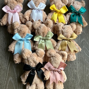 May include: A collection of nine small teddy bears crafted from tan, textured fabric. Each bear features a smooth satin ribbon tied in a bow around its neck, with colors including light pink, white, yellow, dark green, light blue, sage, gold, black, and dusty rose.