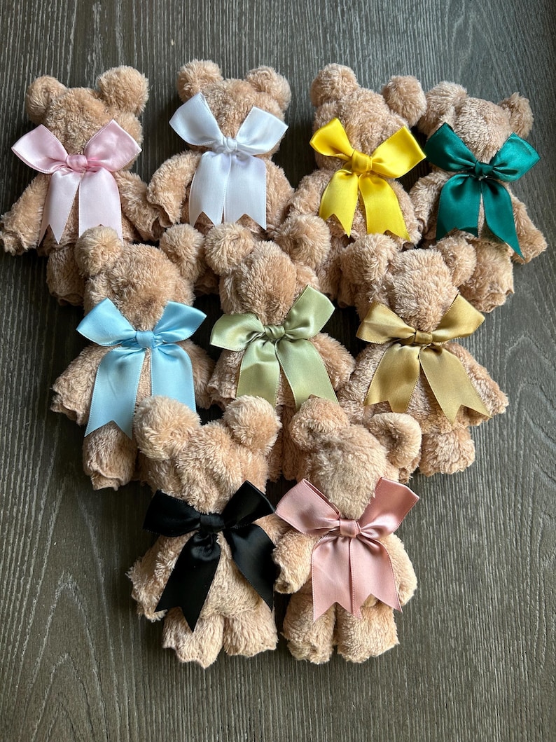 May include: A collection of nine small teddy bears crafted from tan, textured fabric. Each bear features a smooth satin ribbon tied in a bow around its neck, with colors including light pink, white, yellow, dark green, light blue, sage, gold, black, and dusty rose.