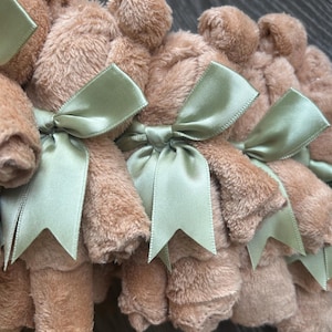 May include: A collection of small, tan, plush fabric bears, each rolled and tied with a sage green satin ribbon. The bears feature a soft, fuzzy texture and are arranged in a row, showcasing the uniform light brown color and the tied bows.