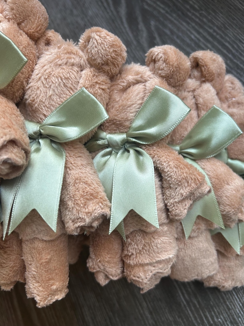 May include: A collection of small, tan, plush fabric bears, each rolled and tied with a sage green satin ribbon. The bears feature a soft, fuzzy texture and are arranged in a row, showcasing the uniform light brown color and the tied bows.