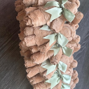 May include: A stack of multiple tan-colored, plush, rolled washcloths. Each individual washcloth is secured with a light sage green satin ribbon tied into a bow. The texture of the fabric appears soft and fuzzy, and the items are arranged in a vertical column.