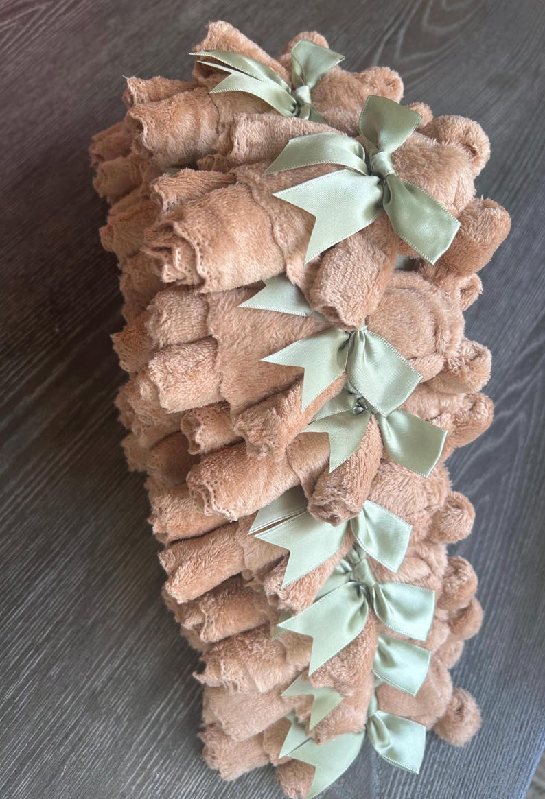 May include: A stack of multiple tan-colored, plush, rolled washcloths. Each individual washcloth is secured with a light sage green satin ribbon tied into a bow. The texture of the fabric appears soft and fuzzy, and the items are arranged in a vertical column.