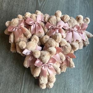 May include: A collection of small, beige plush teddy bears arranged in a cluster. Each bear features a soft, fuzzy texture and is adorned with a shiny, dusty rose-colored satin ribbon tied into a bow around its neck, creating a uniform appearance.