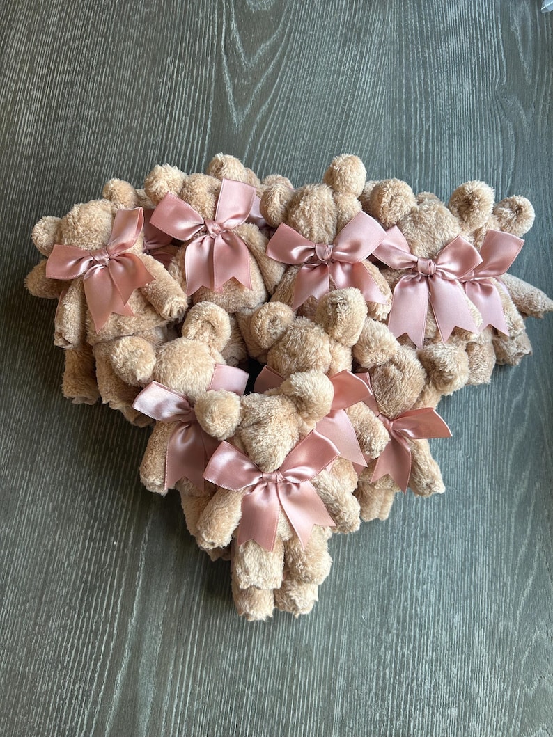 May include: A collection of small, beige plush teddy bears arranged in a cluster. Each bear features a soft, fuzzy texture and is adorned with a shiny, dusty rose-colored satin ribbon tied into a bow around its neck, creating a uniform appearance.