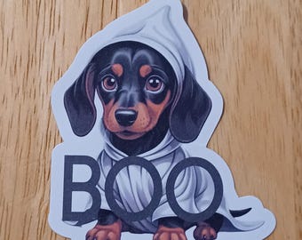 May include: A sticker of a dachshund puppy in a white ghost costume. The puppy has black and brown fur and large brown eyes. The word "BOO" is printed in large black letters below the puppy.