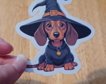 May include: A sticker of a cartoon dachshund wearing a black witch's hat with a brown buckle and a black sweater. The dog has brown fur and large eyes. The sticker has a white border and a die-cut edge.