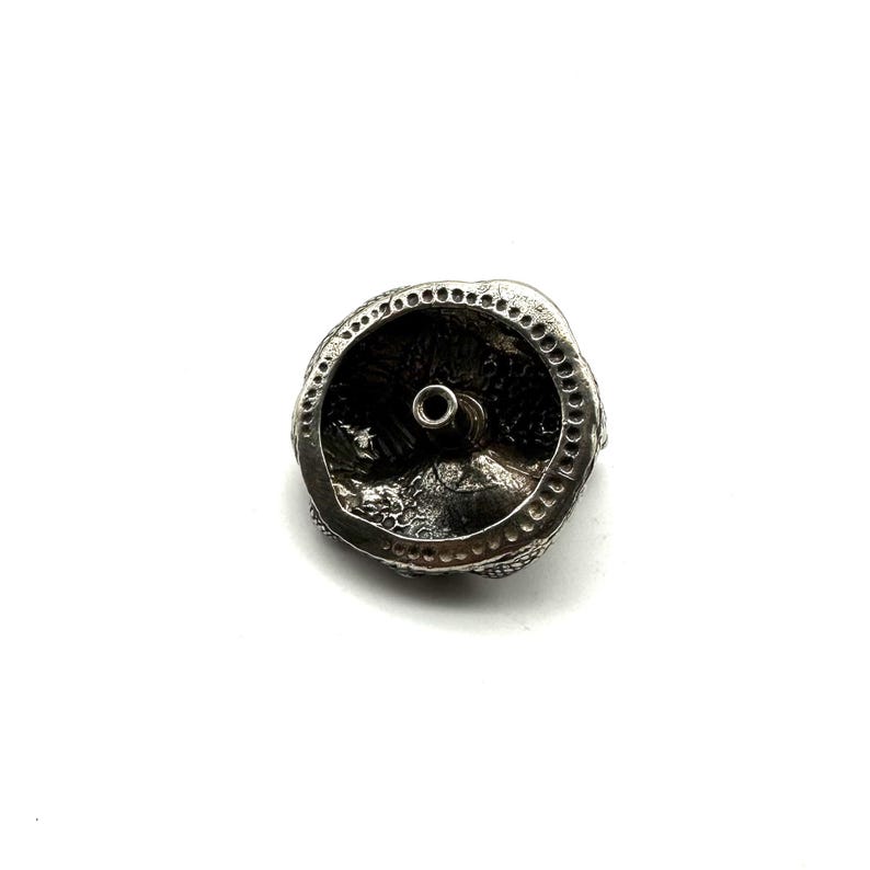 May include: A detailed, silver-coloured metal bead with a central hole. The bead has a textured surface and a decorative, circular design around the edge. The bead is set against a white background.