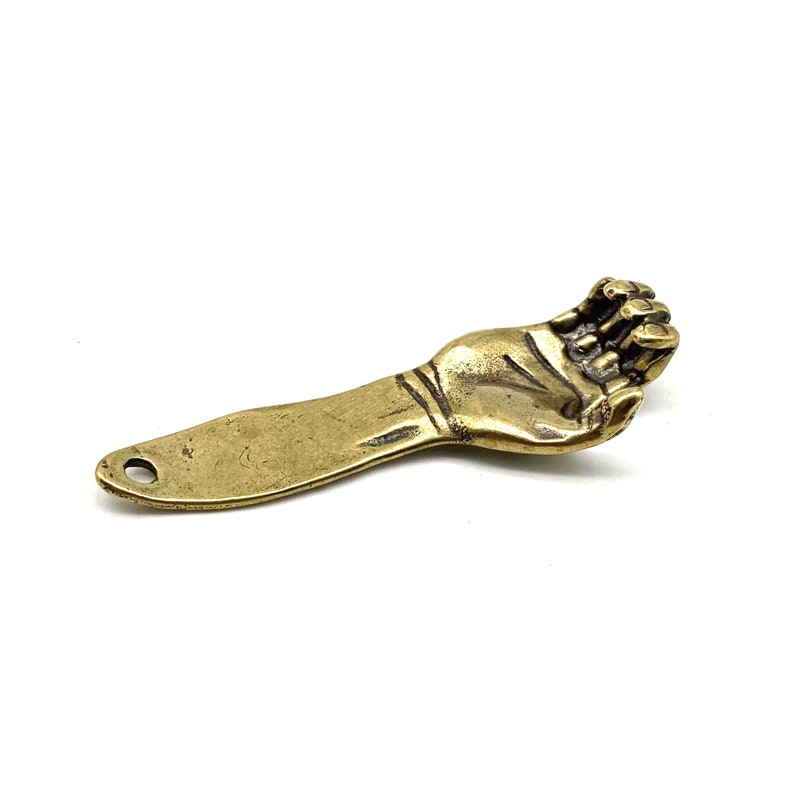 May include: A brass bottle opener shaped like a hand with the fingers curled into a fist. The opener has a hole at the bottom of the handle.