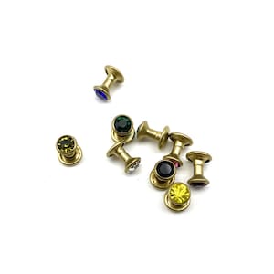 May include: Assortment of gold-toned metal button-like embellishments with various colored rhinestones. The rhinestones are green, blue, red, yellow, black, and clear.