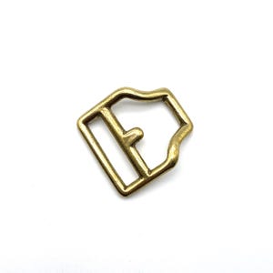 May include: A gold-toned metal buckle with a rectangular shape and a single, centered opening. The buckle is designed for use with straps or belts.