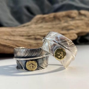 May include: Two silver rings with feather designs. Each ring has a detailed feather pattern and a small, round gold-coloured emblem with a bird silhouette. The rings are displayed on a light surface.