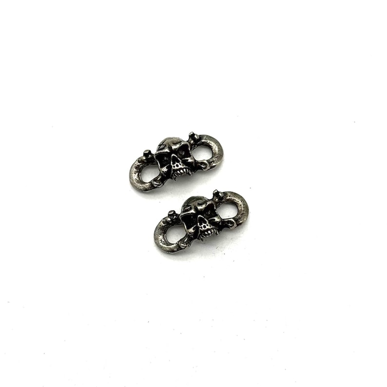 May include: Two silver-toned skull-shaped connectors. Each connector features a detailed skull design with open eye sockets and a grinning mouth. The connectors have a loop on each side, suggesting they can be used to connect other items. The background is white.