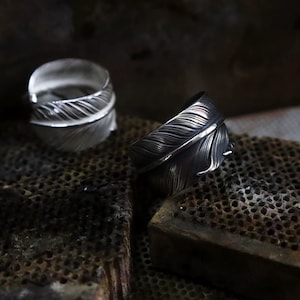 May include: Two silver rings featuring a feather design. The rings are intricately detailed with textured lines, mimicking the barbs of a feather. They are presented on a textured, dark surface, with one ring in the foreground and another in the background.