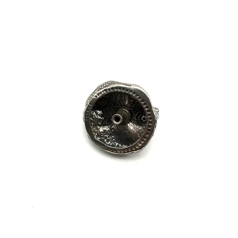 May include: A silver bottle stopper with a detailed, textured design. The stopper has a circular base with a central hole and a decorative top. The design appears to be of a mythical creature or animal.