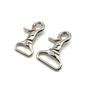 May include: Two silver-colored swivel snap hooks. Each hook has a spring-loaded clasp and a D-ring base. The hooks are made of shiny metal and are set against a white background.
