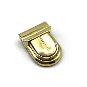 Pode incluir: Um fecho de metal dourado com uma parte superior arredondada e arqueada e uma parte inferior retangular com dobradiças. O fecho tem um acabamento liso e polido.