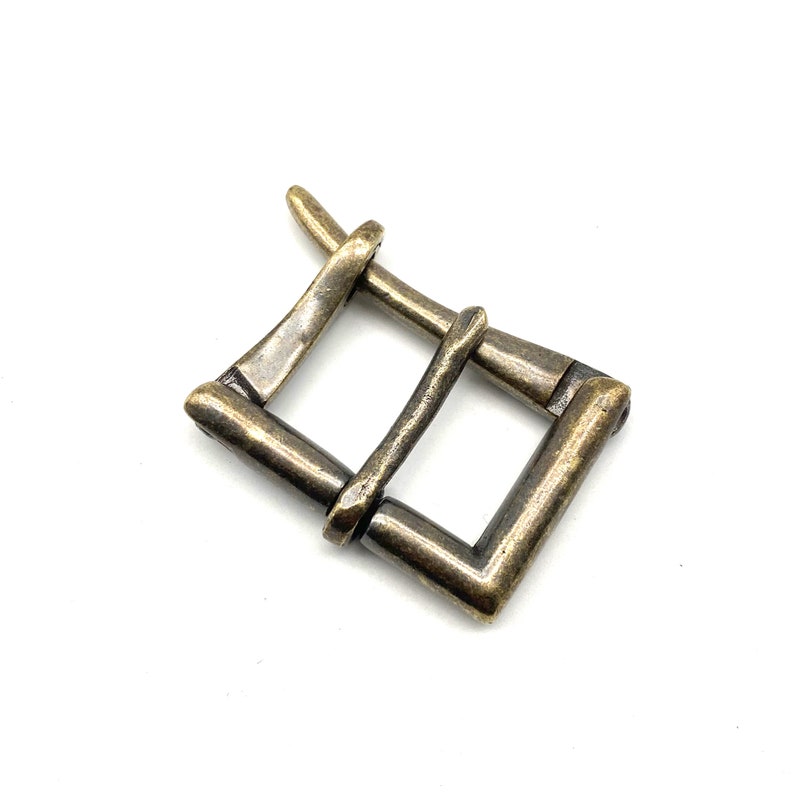 May include: A gold-toned metal buckle with a rectangular shape and a unique, intricate design. The buckle has a slightly aged, antique look.