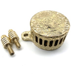 May include: A gold-colored, intricately engraved metal object with the text "SIC PARVIS MAGNA". The object has a round, domed shape with a handle and two smaller, threaded components. The design features ornate patterns.