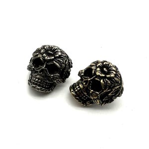 Op de afbeelding: Twee zilveren schedelcharms met bloemdetails. De schedel links is zilver, de schedel rechts is goud.