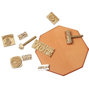 May include: A set of brass leather stamping tools, including a screw, a rectangular stamp with the text "EIGTE" and "DEBUT 1883", a square stamp with a Christmas tree design, a round stamp with a floral design, a rectangular stamp with the text "ATUW", a rectangular stamp with the text "Best Wishes To You", and a rectangular stamp with a floral design.