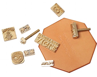 May include: A set of brass leather stamping tools, including a screw, a rectangular stamp with the text "EIGTE" and "DEBUT 1883", a square stamp with a Christmas tree design, a round stamp with a floral design, a rectangular stamp with the text "ATUW", a rectangular stamp with the text "Best Wishes To You", and a rectangular stamp with a floral design.