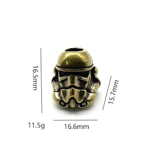 May include: A gold-coloured metal charm in the shape of a Star Wars Stormtrooper helmet. The charm is 16.6 mm wide and 15.7 mm tall. It weighs 11.5 grams.