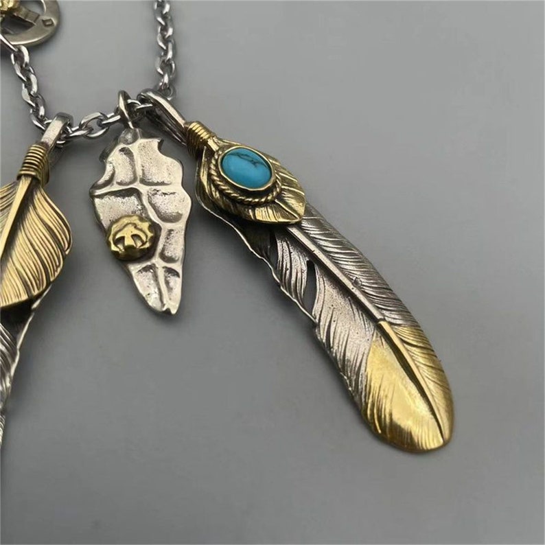May include: A silver and gold necklace with three feather pendants. The center feather has a turquoise stone set in a gold bezel. The necklace is on a silver chain.