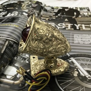 May include: Gold motorcycle tail light with a red lens and intricate floral engravings. The light is mounted on a gold bracket with wiring attached.