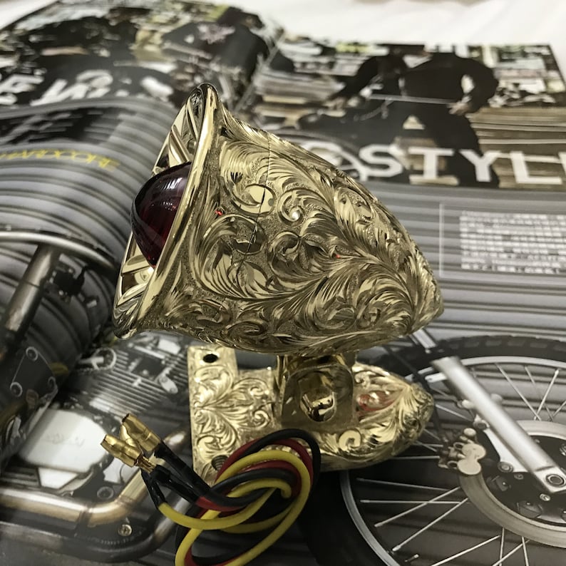May include: Gold motorcycle tail light with a red lens and intricate floral engravings. The light is mounted on a gold bracket with wiring attached.