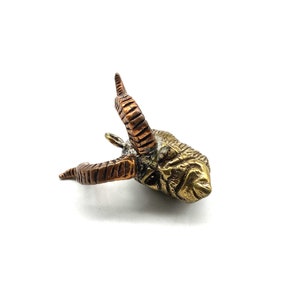 May include: A detailed brass sculpture of a ram's head, featuring textured horns in shades of copper and a golden body. The sculpture is set against a white background, highlighting the intricate details of the animal's face.