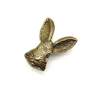 Pode incluir: Uma escultura de cabeça de coelho de metal dourado com detalhes precisos. As orelhas do coelho são pontiagudas e a escultura está voltada para a frente.