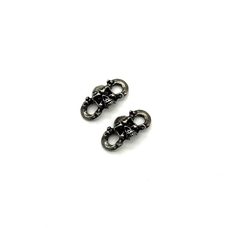 May include: Two small, silver-toned metal charms, each featuring a skull design. The skulls are detailed with visible teeth and eye sockets, connected by a loop. The charms are set against a plain white background.